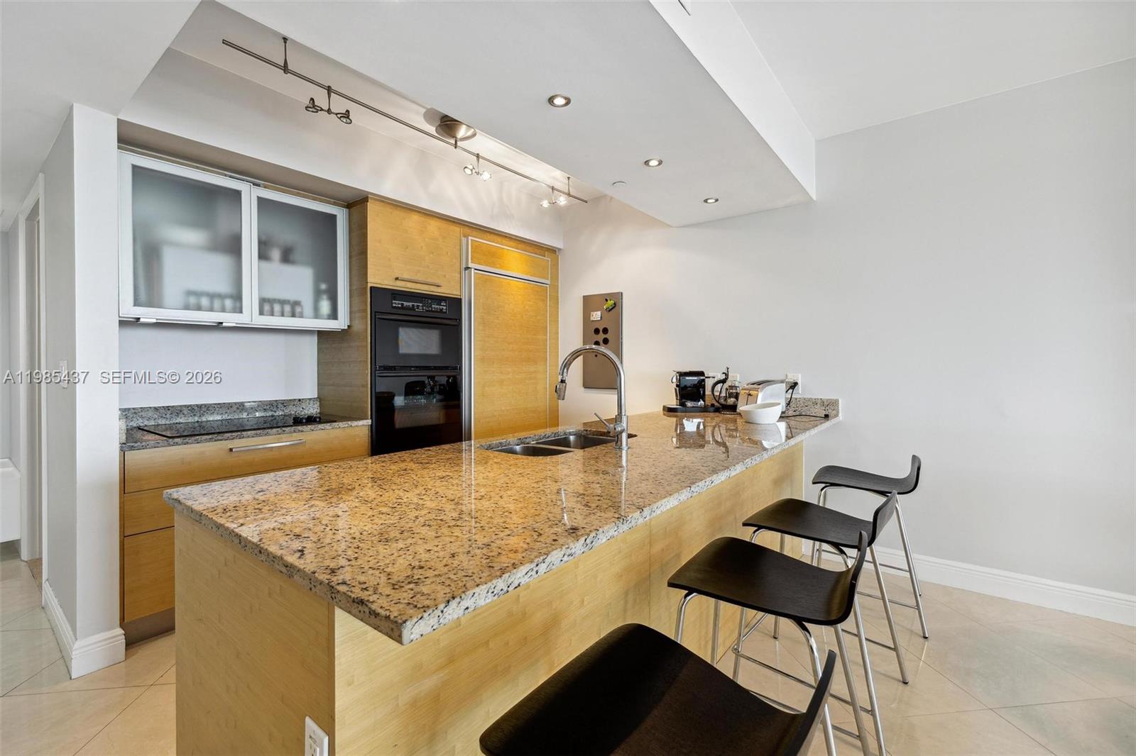 A kitchen features light wood cabinetry, granite countertops, a central island, and three black stools — 1830 S Ocean Dr # 2703, Hallandale Beach FL 33009, HALLANDALE BEACH, FL