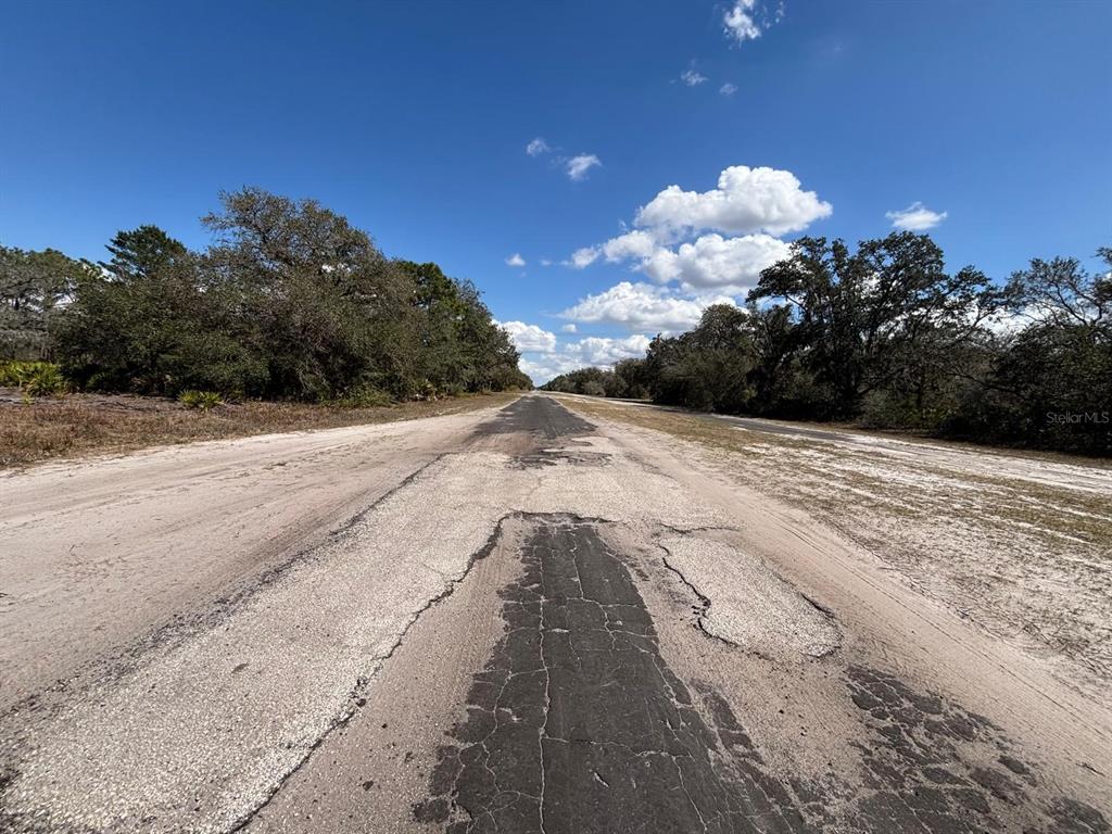 A weathered, narrow asphalt road stretches through a rural landscape with trees and sandy shoulders — 1006 PALM AVENUE, INDIAN LAKE ESTATES, FL