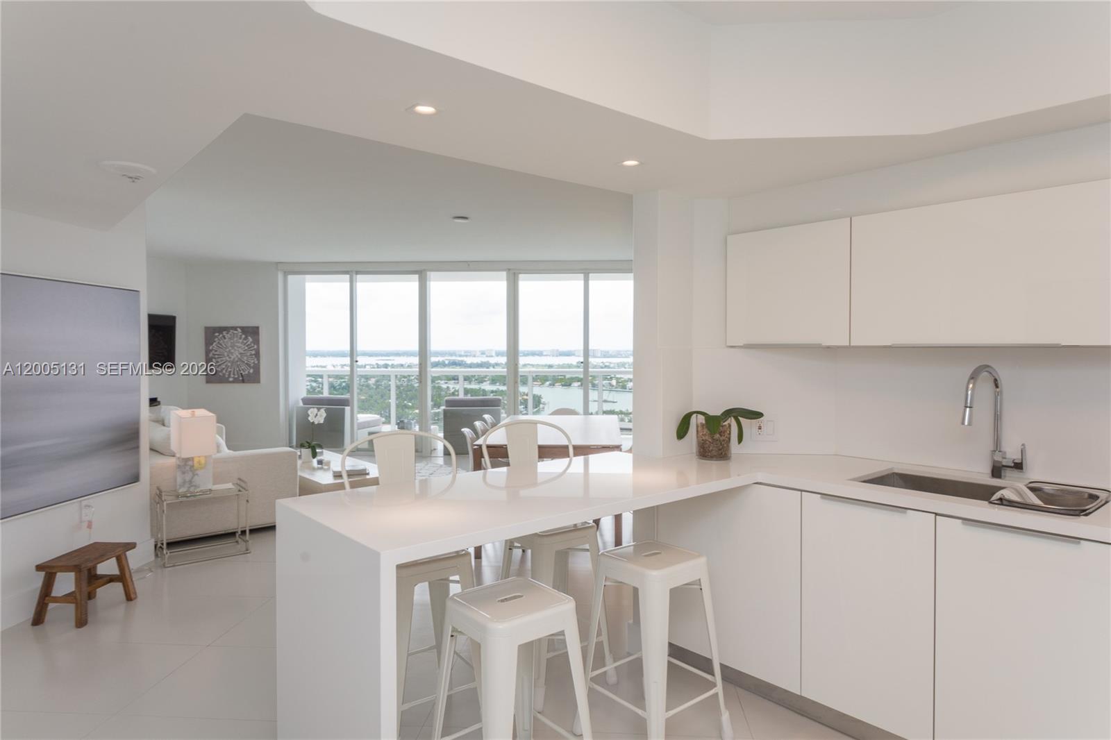 A modern white kitchen features a breakfast bar with stools overlooking a bright living area — 6301 Collins Ave # 3006, Miami Beach FL 33141, MIAMI BEACH, FL