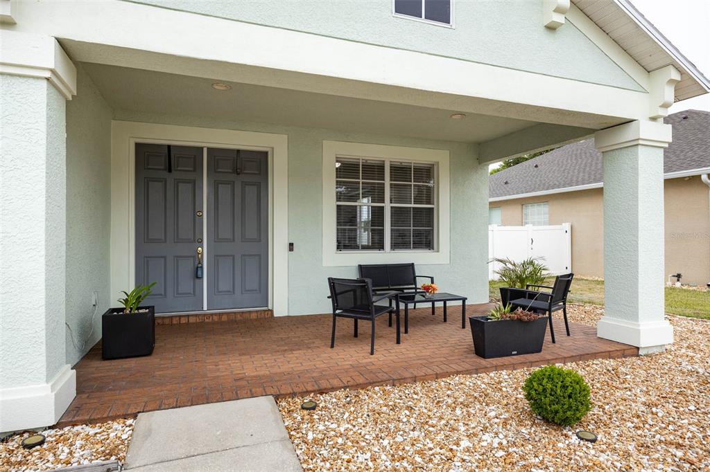A covered front porch features a brick floor, double gray doors, and outdoor patio furniture — 4215 MAPLEHURST WAY, SPRING HILL, FL