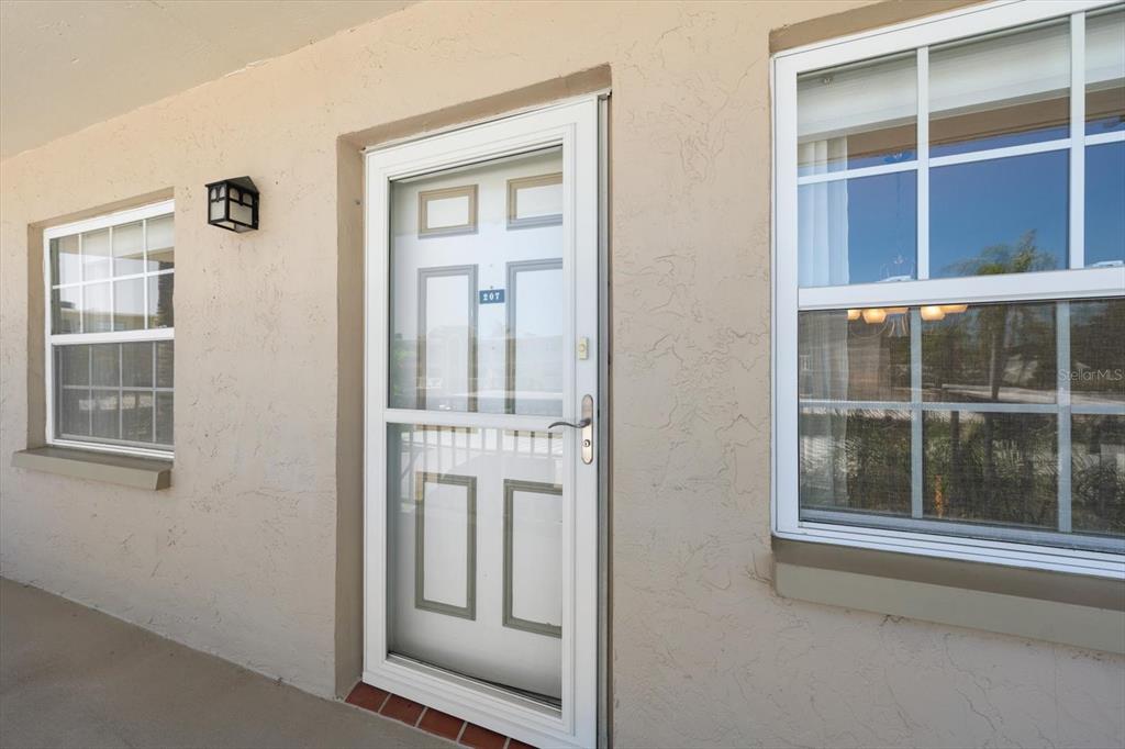 A white storm door with the number 207 is flanked by two windows on a stucco wall — 948 VIRGINIA STREET UNIT 207, DUNEDIN, FL