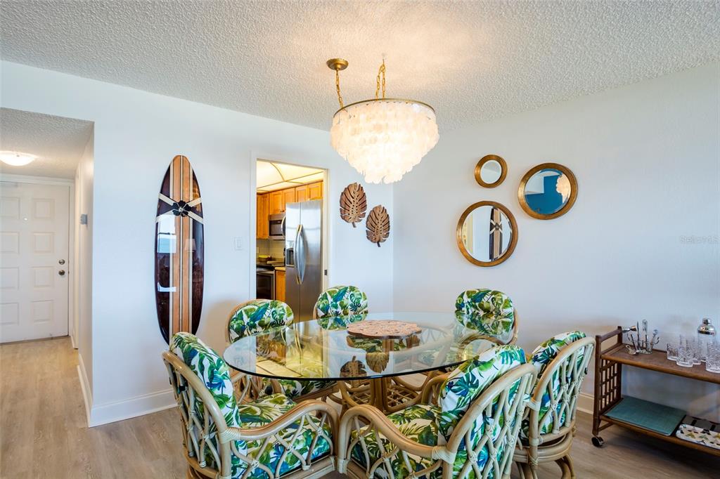 A dining room features a glass-topped rattan table, tropical-print chairs, a surfboard, and a chandelier — 1230 GULF BOULEVARD UNIT 903, CLEARWATER BEACH, FL