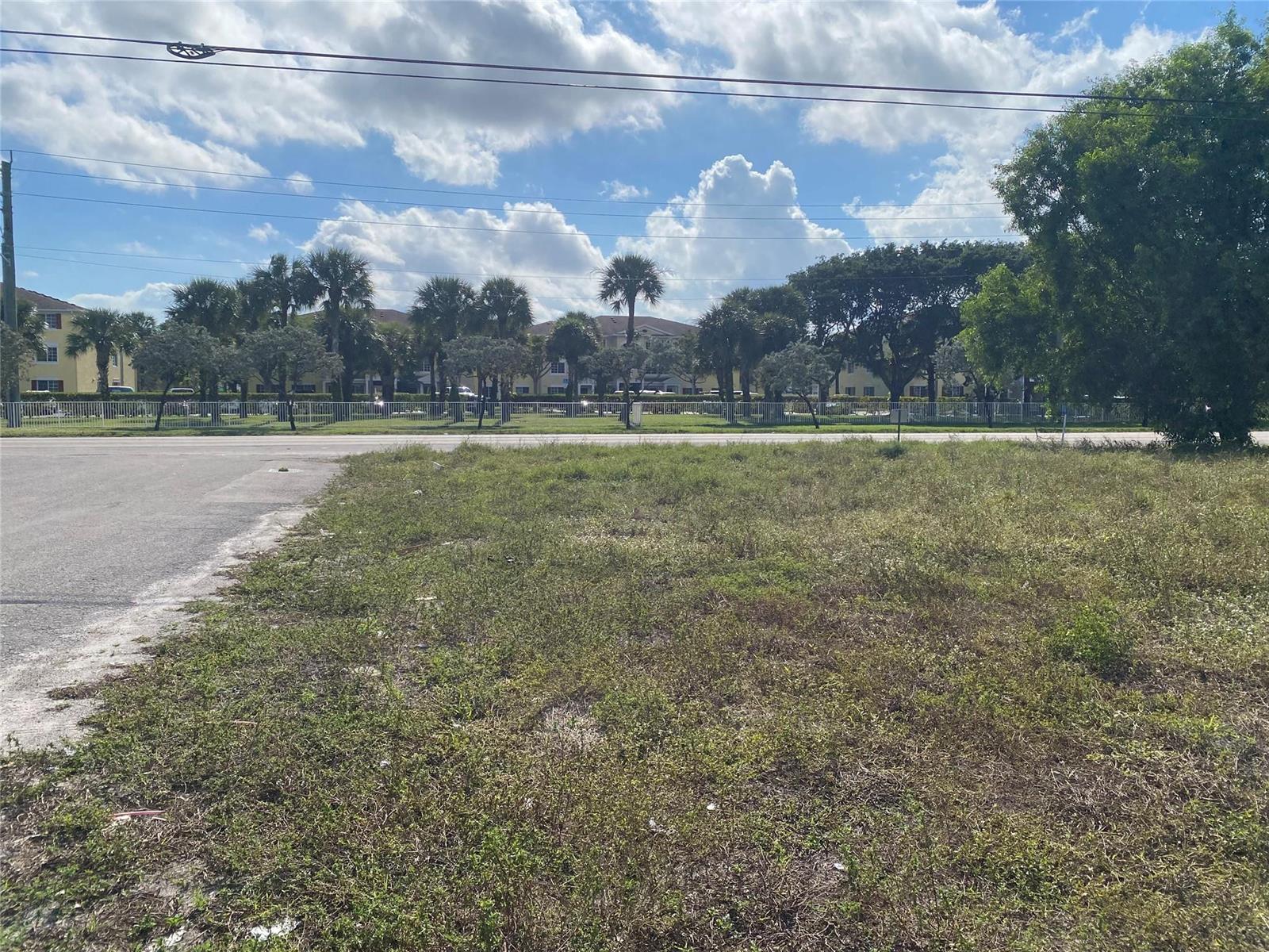 A vacant, grassy lot sits beside a paved road with apartment buildings in the background — Nw 19 NW 19th Avenue, Pompano Beach FL 33069, POMPANO BEACH, FL