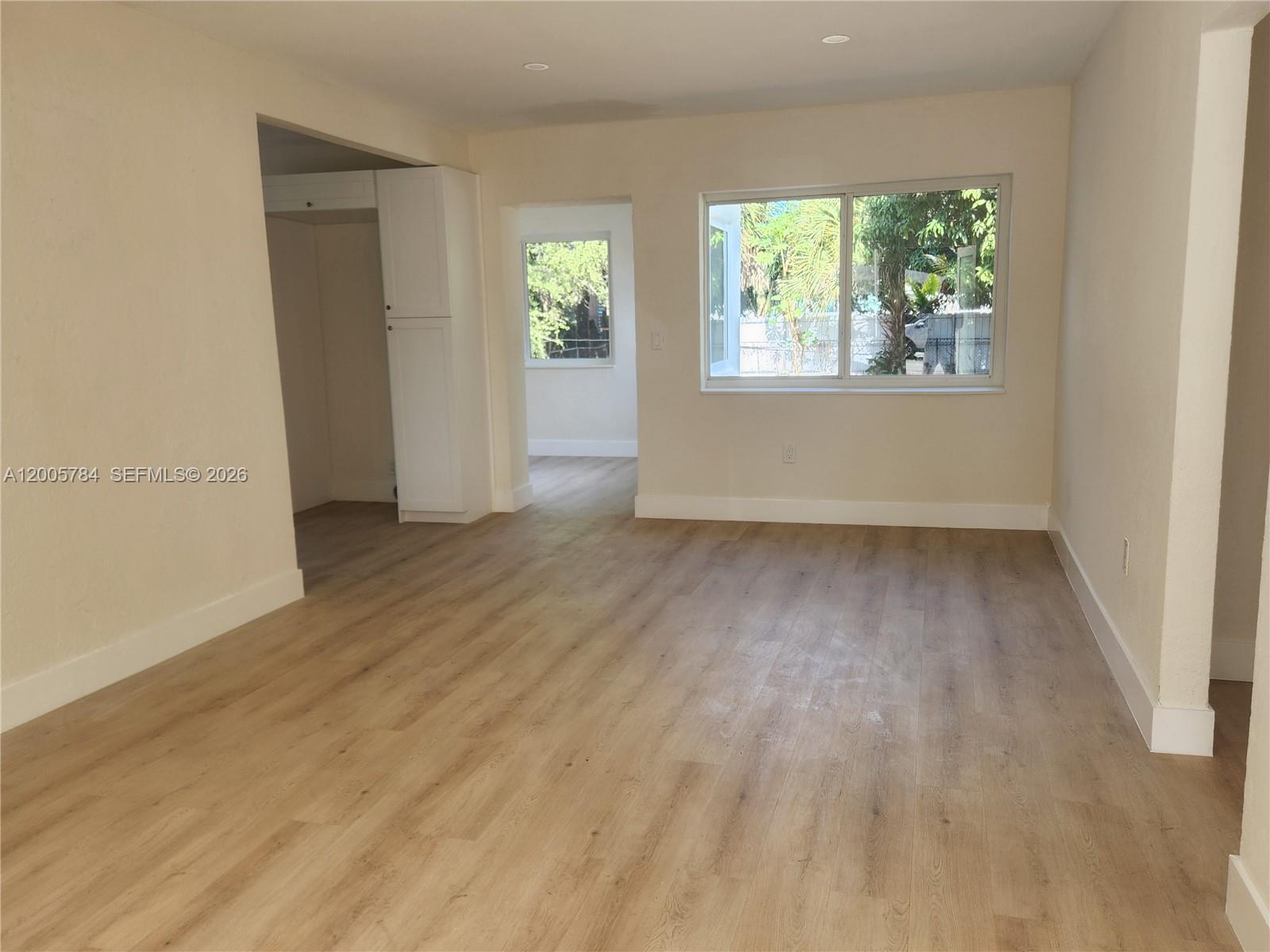 An empty room features light wood-look flooring, beige walls, and a window overlooking greenery — 815 NE 134th St, North Miami FL 33161, NORTH MIAMI, FL