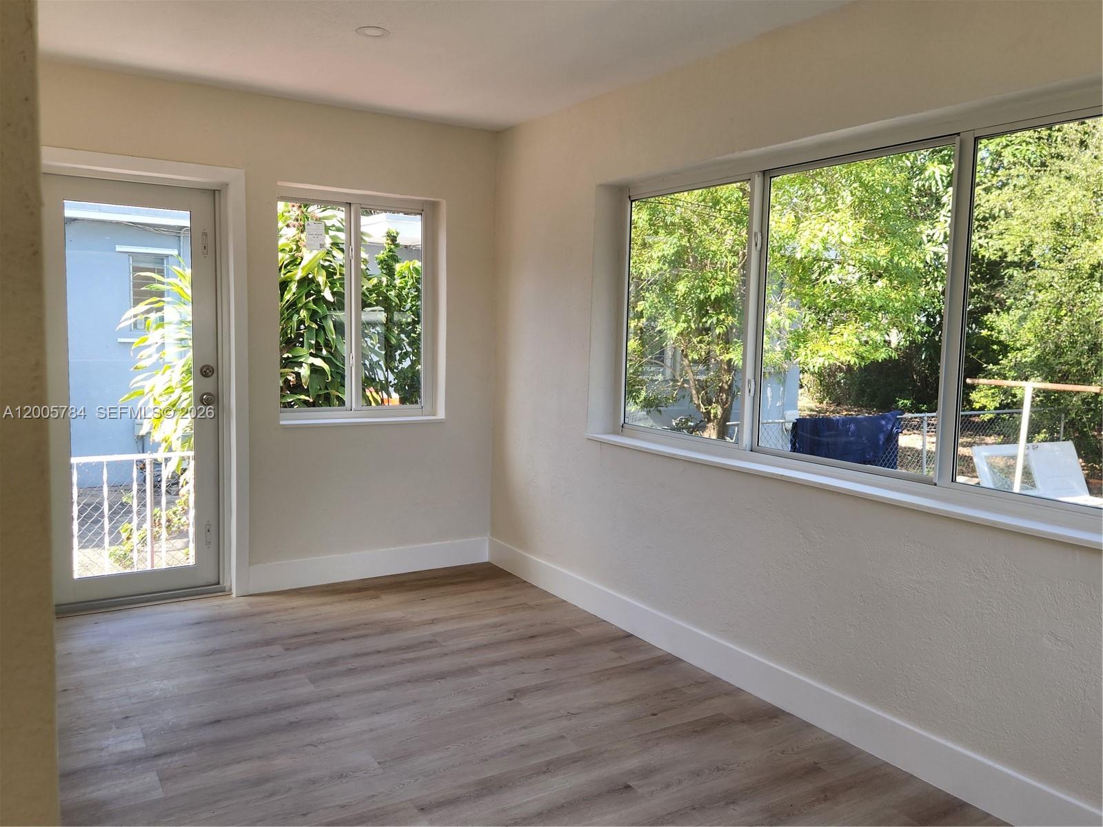 A bright room features light wood-look flooring, beige walls, and large windows overlooking trees — 815 NE 134th St, North Miami FL 33161, NORTH MIAMI, FL