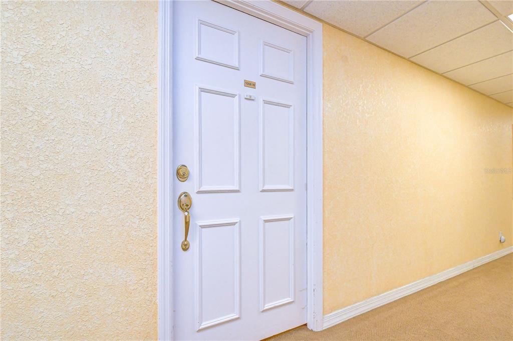 A white six-panel door with gold hardware is set into a textured beige wall — 400 64TH AVENUE UNIT 1006, ST PETE BEACH, FL
