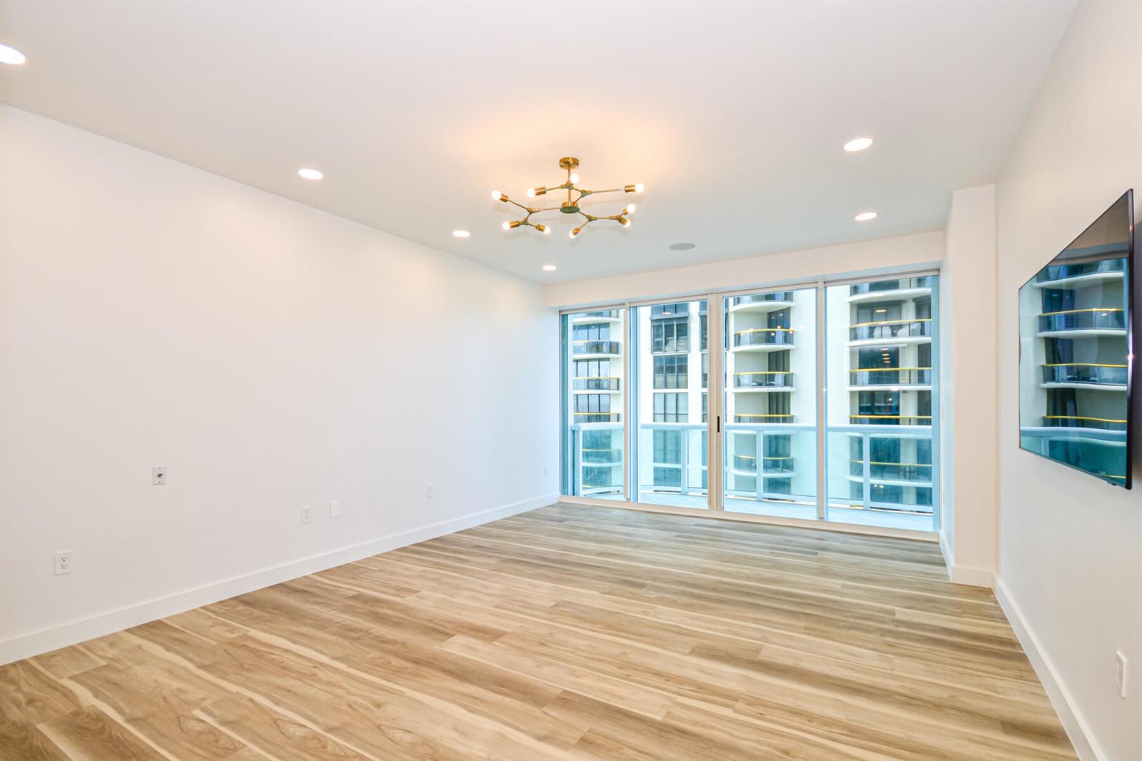 An empty living room with light wood flooring, a modern chandelier, and floor-to-ceiling windows — 10101 Collins Avenue # 7d, Bal Harbour FL 33154, BAL HARBOUR, FL