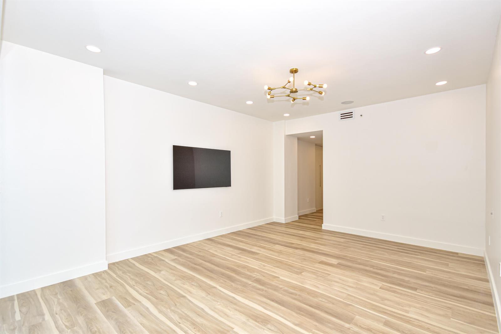 An empty room features light wood-look flooring, white walls, a mounted television, and a chandelier — 10101 Collins Avenue # 7d, Bal Harbour FL 33154, BAL HARBOUR, FL