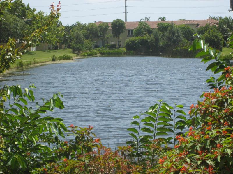 A view of a calm pond surrounded by lush greenery and a multi-story apartment building — 138 Monterey Bay Drive, Boynton Beach FL 33426, BOYNTON BEACH, FL