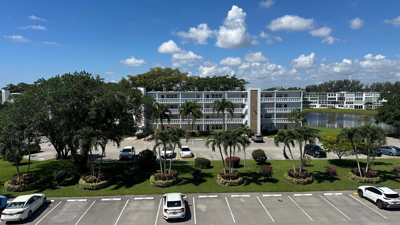 An exterior view of a multi-story apartment building with a parking lot and palm trees — 1044 Cambridge C, Deerfield Beach FL 33442, DEERFIELD BEACH, FL