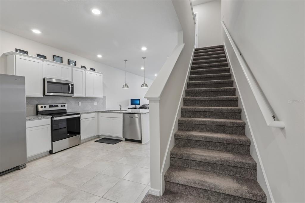 A kitchen with white cabinets and stainless steel appliances sits beside a carpeted staircase — 31337 AMBERVIEW BEND, WESLEY CHAPEL, FL