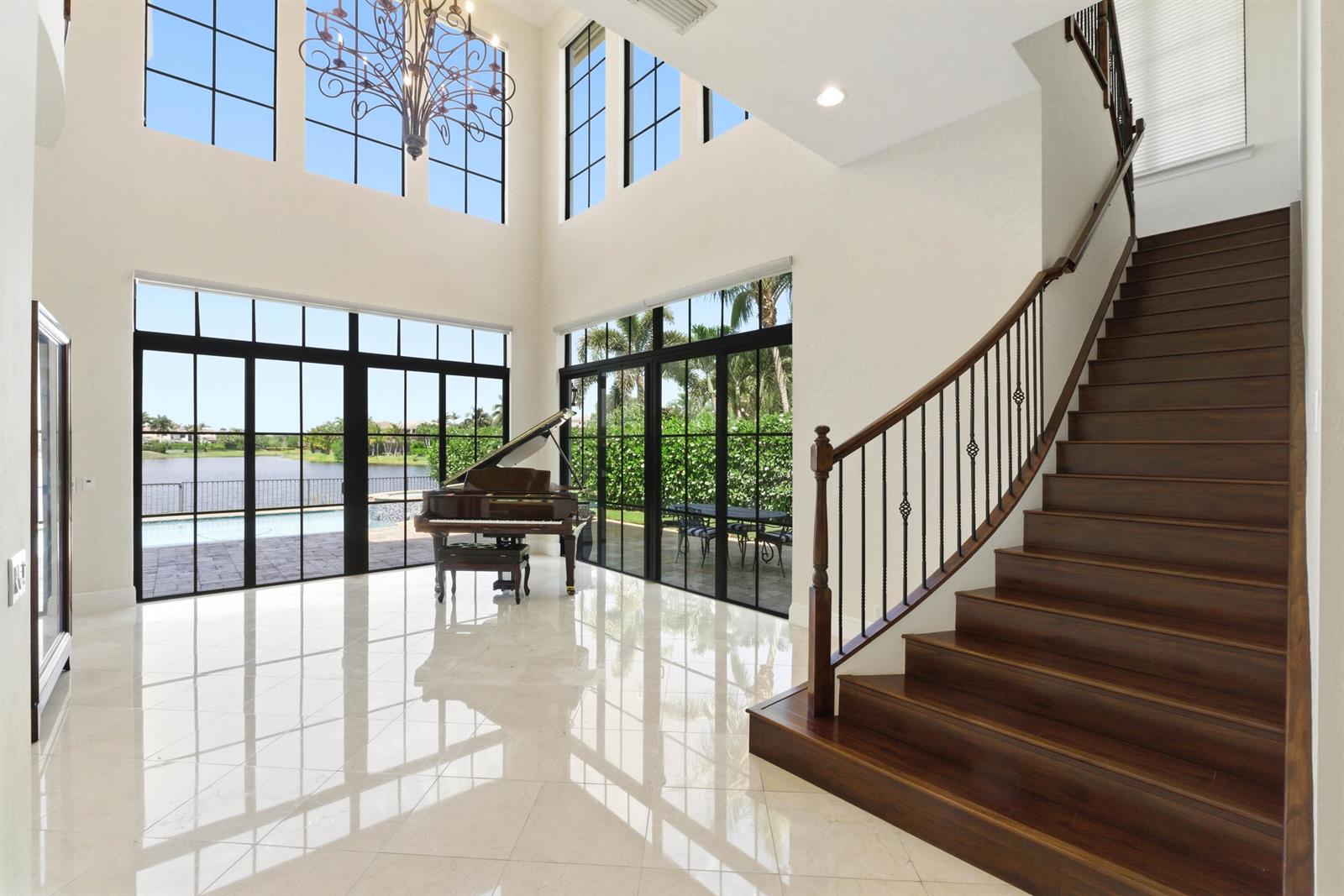 A grand foyer with a grand piano, marble floors, a curved staircase, and floor-to-ceiling windows — 8289 Emerald Avenue, Parkland FL 33076, PARKLAND, FL