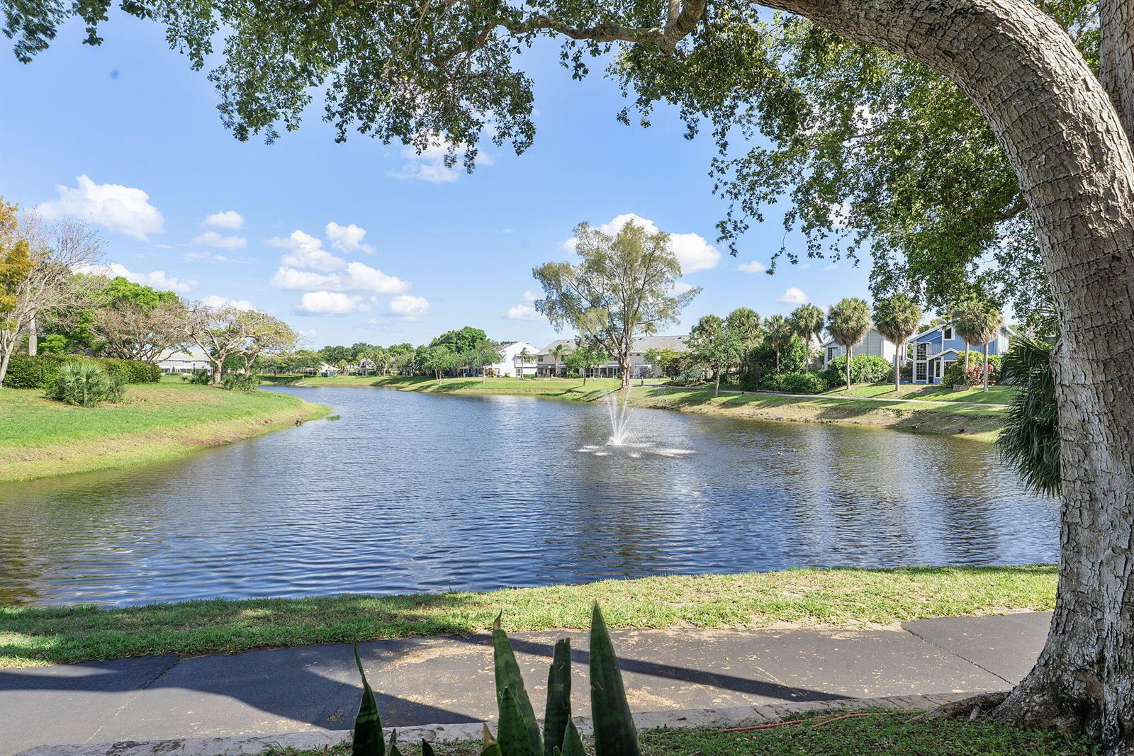 A view of a lake with a fountain, surrounded by grass, trees, and residential buildings — 5272 Buckhead Circle # 1010, Boca Raton FL 33486, BOCA RATON, FL