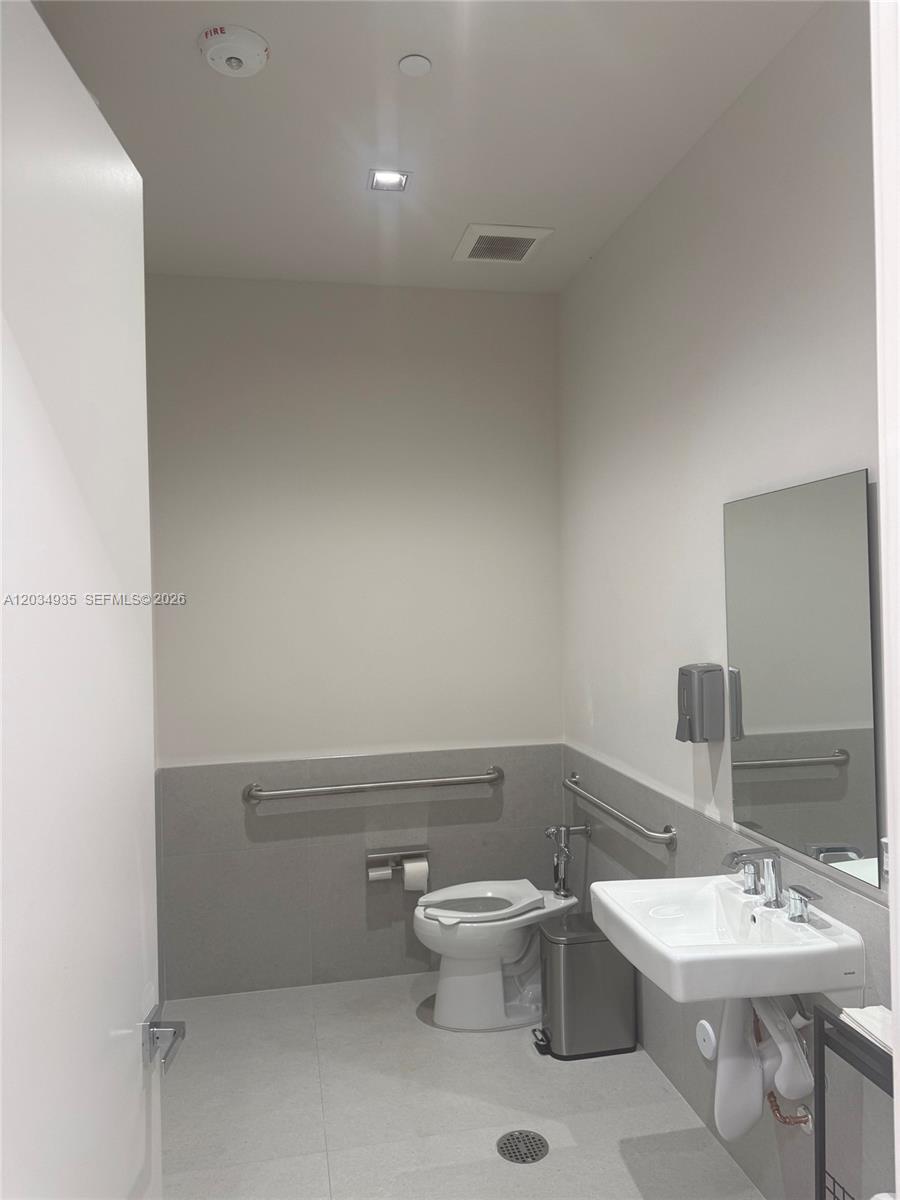A modern, accessible bathroom features a white toilet, wall-mounted sink, and gray tile wainscoting — SWEETWATER, FL