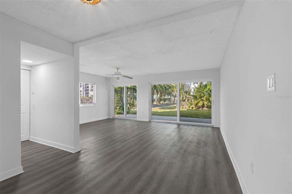 An empty living room features gray wood-look flooring, white walls, and large sliding glass doors — 1451 GULF BOULEVARD UNIT 101, CLEARWATER BEACH, FL
