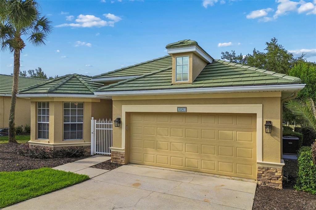 A tan house with a green tile roof and a two-car garage under a blue sky — 13128 PALMILLA CIRCLE, DADE CITY, FL