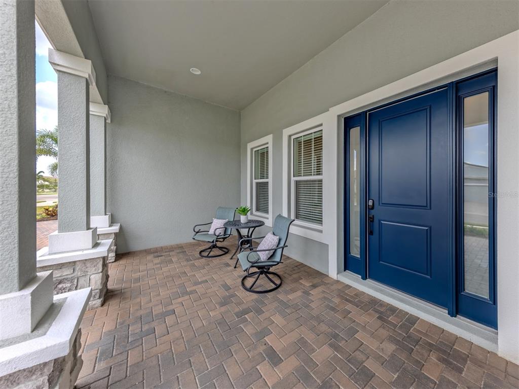 A covered front porch features paver flooring, two chairs, a small table, and a blue door — 19916 BRIDGETOWN LOOP, VENICE, FL