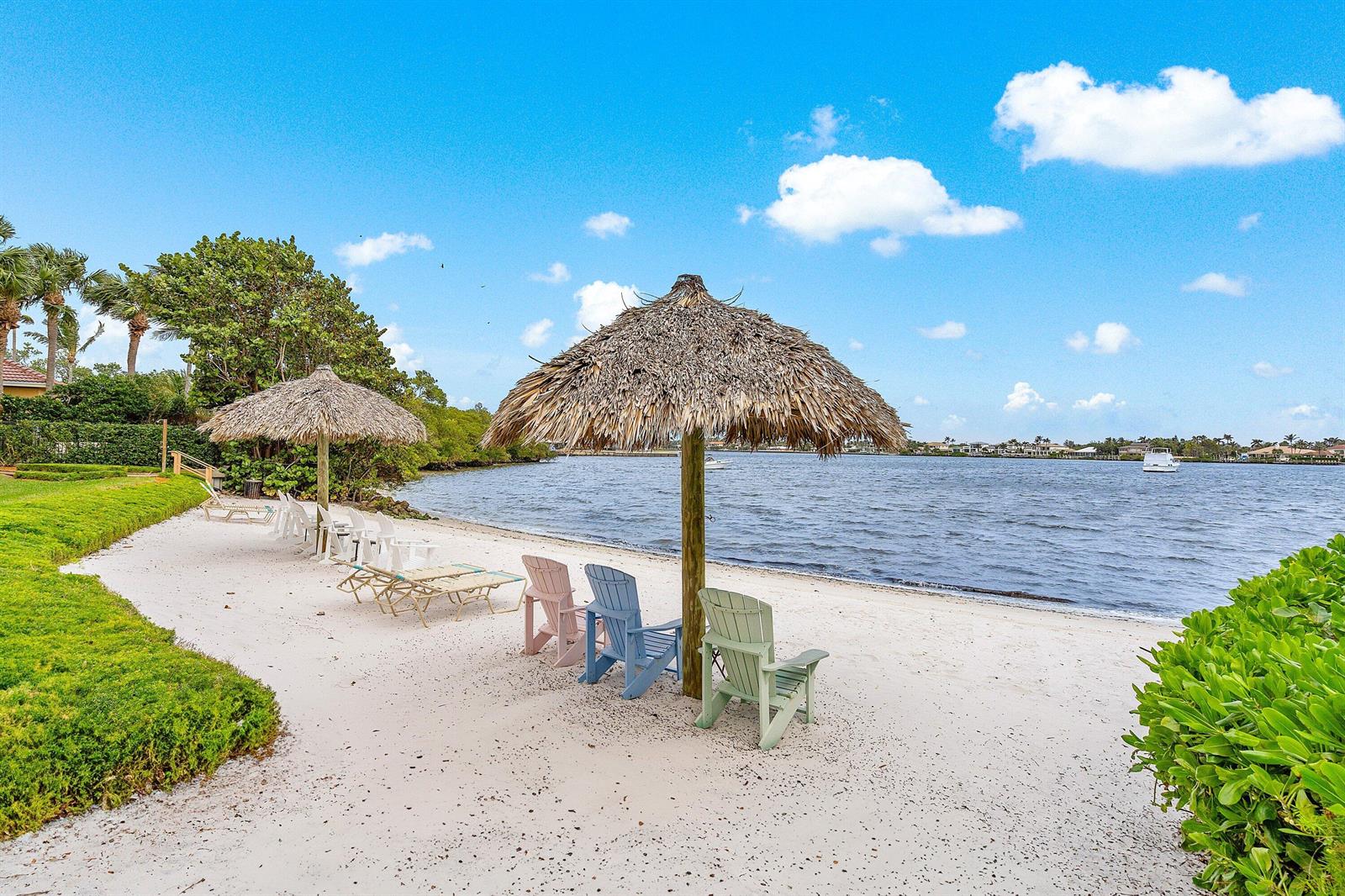 A sandy beach area features two thatched umbrellas and several colorful Adirondack chairs by water — 900 Scotia Drive # 102, Hypoluxo FL 33462, HYPOLUXO, FL
