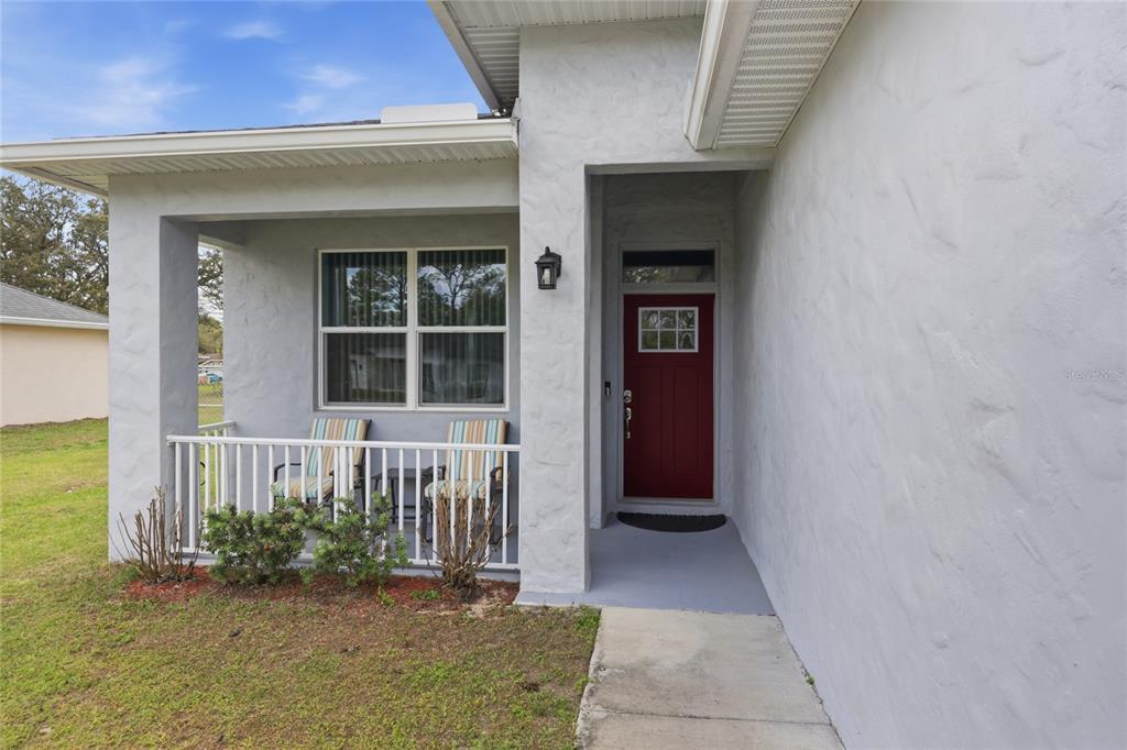 A front porch with a red door, white railing, and two chairs sits beside a lawn — 2118 W HAMPSHIRE BOULEVARD, CITRUS SPRINGS, FL