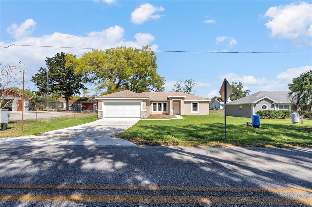 A single-story house with a white garage door and tan exterior sits behind a lawn — 8677 SE 157TH PLACE, SUMMERFIELD, FL