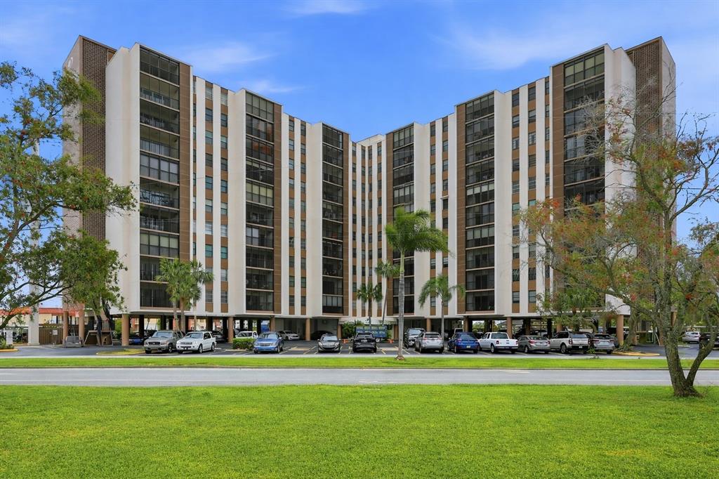 A multi-story apartment building with a parking lot at the base and surrounding trees — 10355 PARADISE BOULEVARD UNIT 608, TREASURE ISLAND, FL
