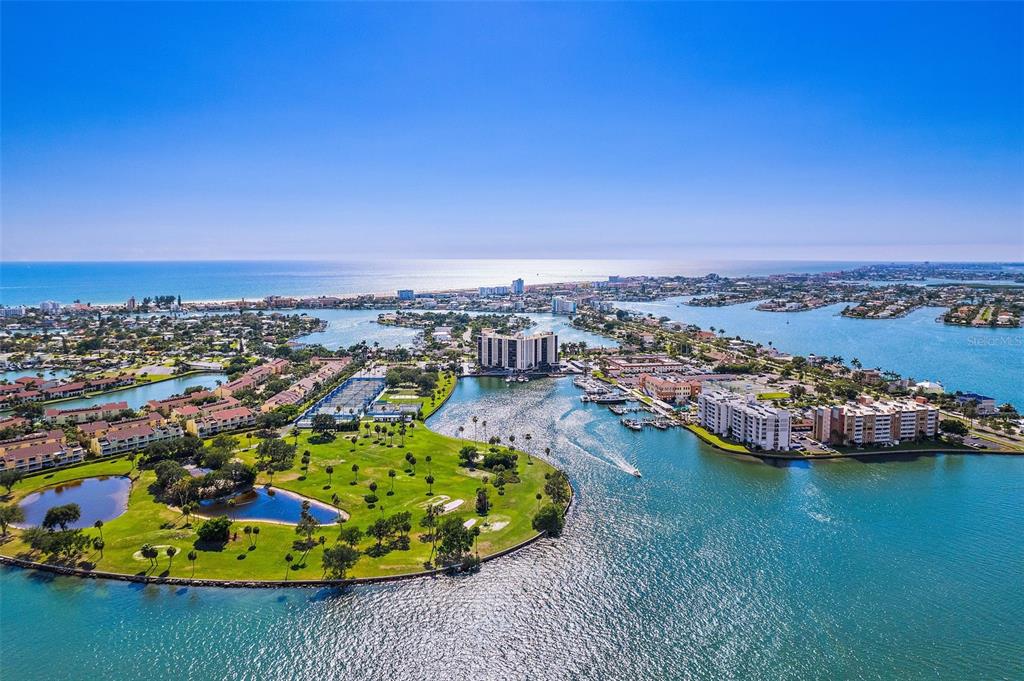 An aerial view shows a coastal community with a golf course, waterways, and residential buildings — 10355 PARADISE BOULEVARD UNIT 608, TREASURE ISLAND, FL