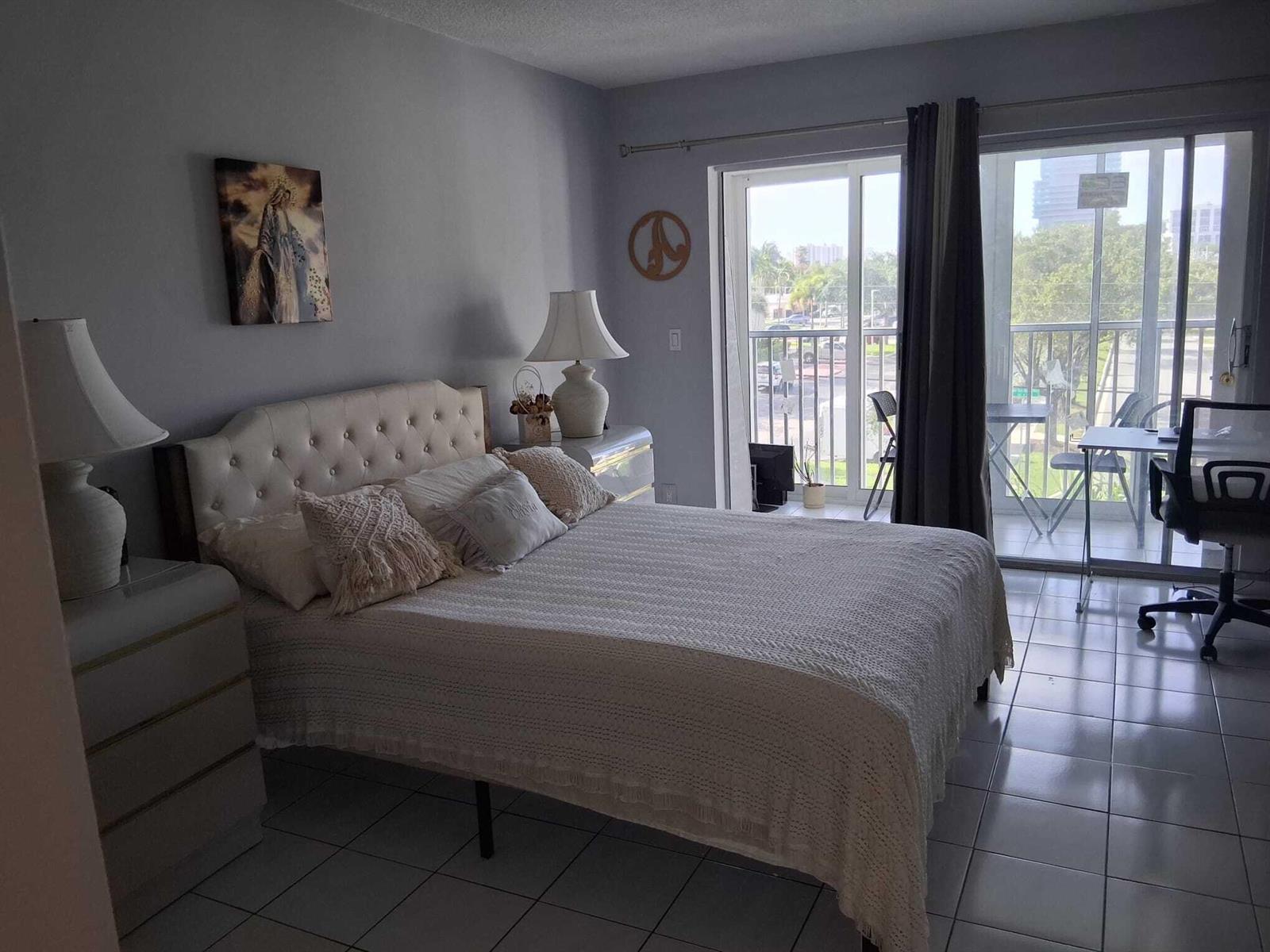 A bedroom with a tufted headboard, tile floors, and a sliding glass door to a balcony — 329 SE 3rd Street # 302s, Hallandale Beach FL 33009, HALLANDALE BEACH, FL