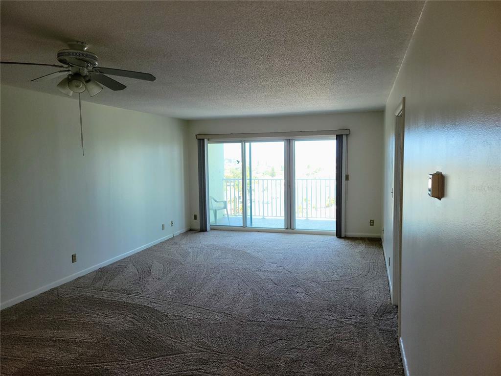 An empty living room features beige carpet, a ceiling fan, and sliding glass balcony doors — 400 64TH AVENUE UNIT 1001 W, ST PETE BEACH, FL