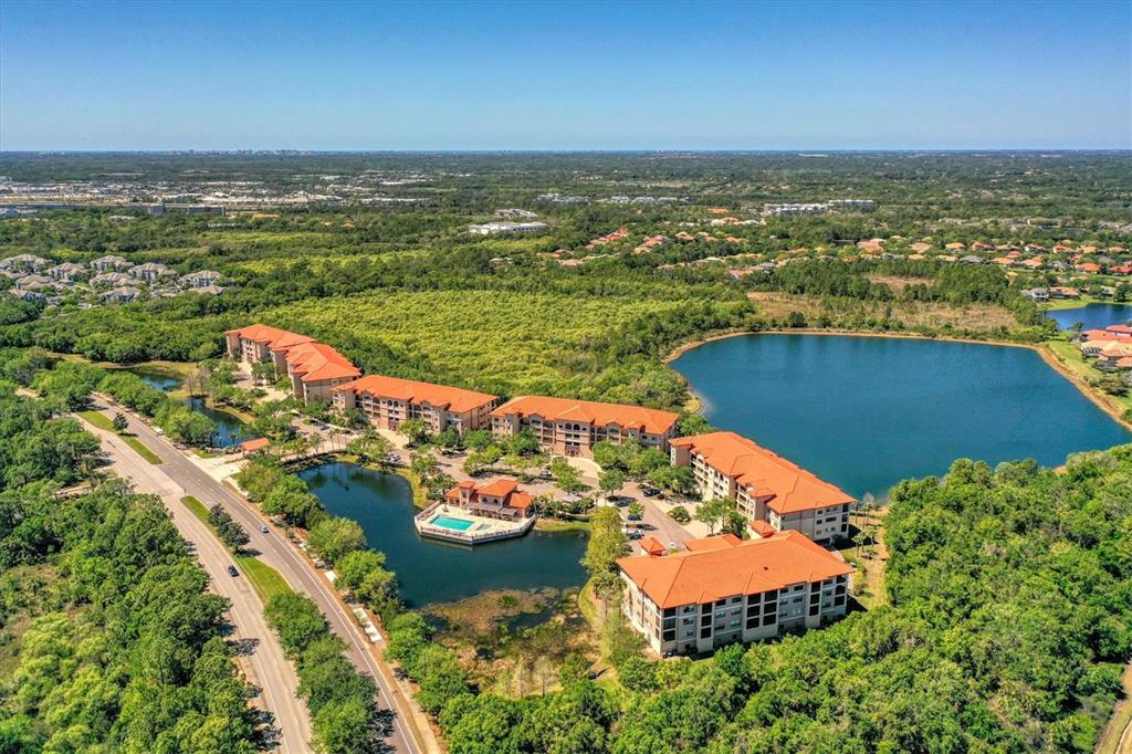 An aerial view of a multi-story apartment complex with red roofs beside a large lake — 7710 LAKE VISTA COURT UNIT 205, LAKEWOOD RANCH, FL