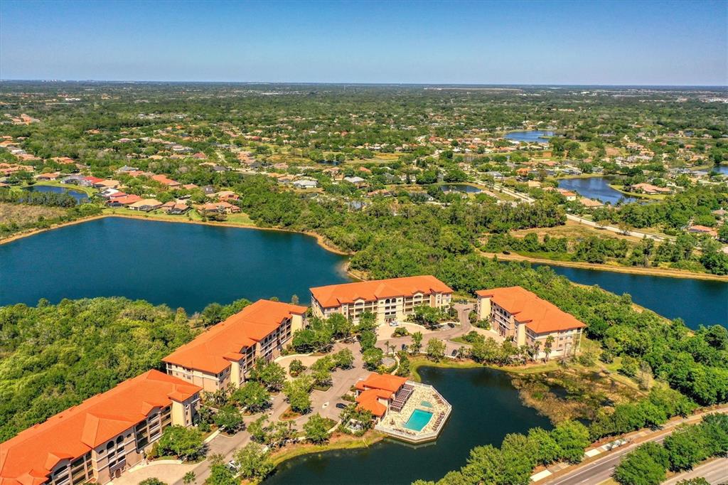 An aerial view shows multi-story apartment buildings with orange roofs surrounded by lakes and trees — 7710 LAKE VISTA COURT UNIT 205, LAKEWOOD RANCH, FL