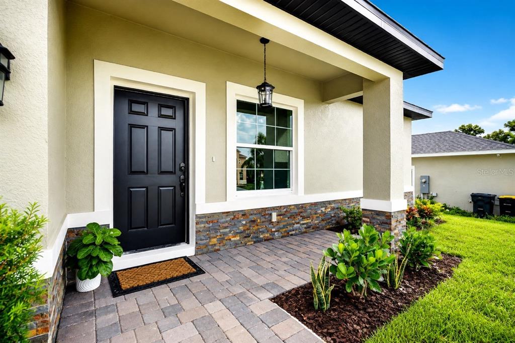 A covered front porch with a black door, stone veneer base, and paver flooring — 1854 SUPERIOR COURT, POINCIANA, FL