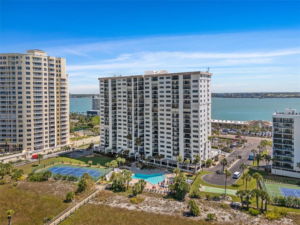 An aerial view shows a tall apartment building with a swimming pool and tennis courts — 1230 GULF BOULEVARD UNIT 303, CLEARWATER BEACH, FL