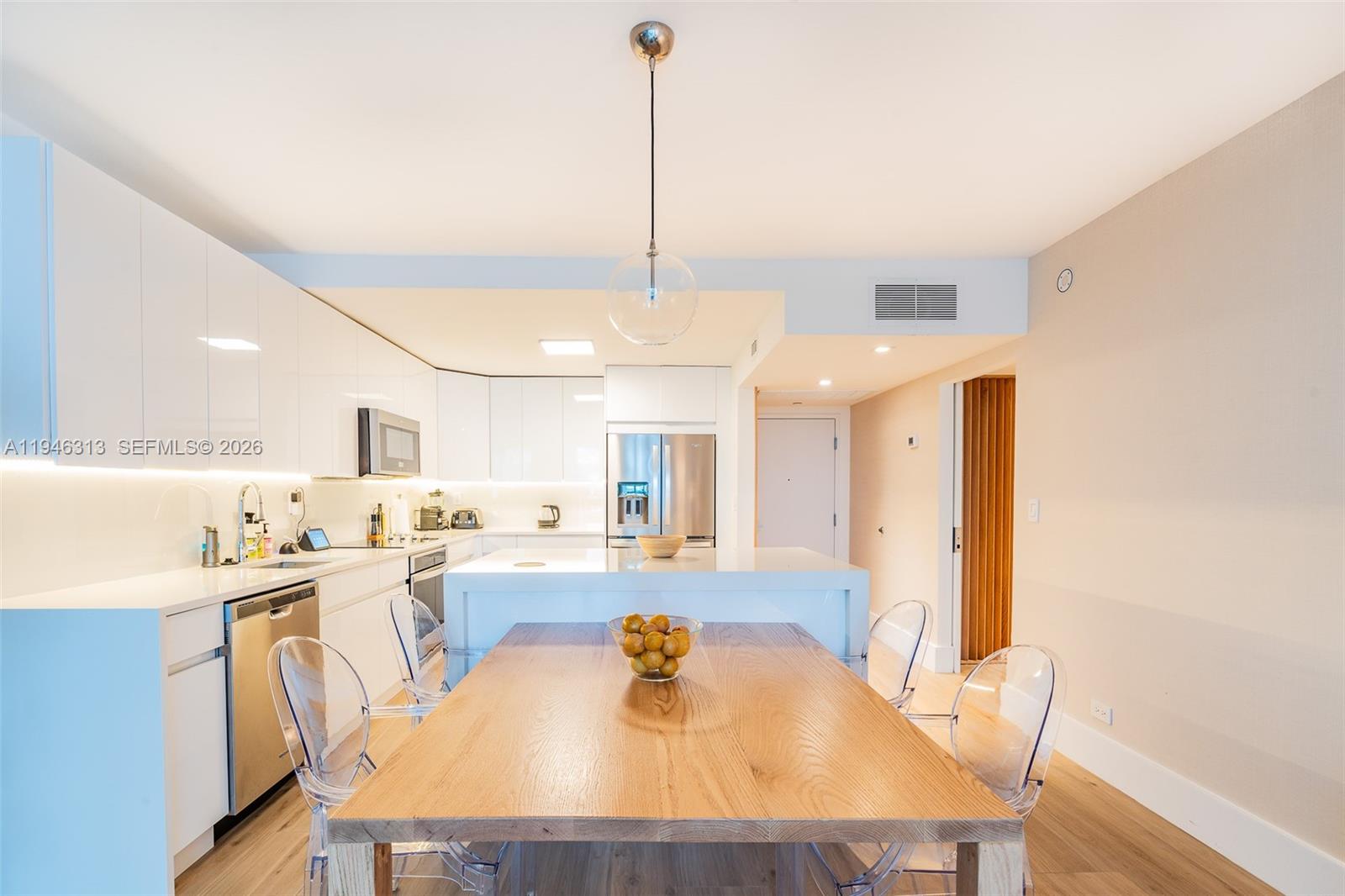 A modern kitchen features white cabinetry, a wooden dining table, and transparent chairs — 2301 Collins Ave # 719, Miami Beach FL 33139, MIAMI BEACH, FL