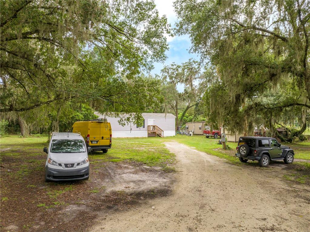 A gravel driveway leads to a light-colored mobile home surrounded by large, moss-draped trees — 36720 EMERALDA ISLAND ROAD, LEESBURG, FL