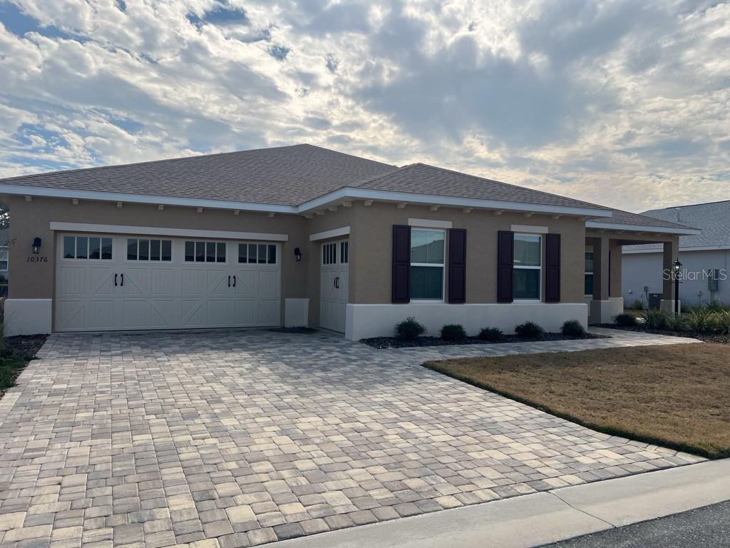 A tan single-story house features a paver driveway, a three-car garage, and dark window shutters — 10376 SW 99 STREET, OCALA, FL