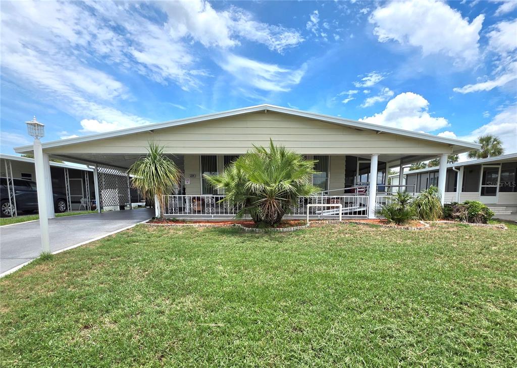 A single-story manufactured home features a large carport, a front porch, and a grassy yard — 5334 COMANCHE STREET, ZEPHYRHILLS, FL