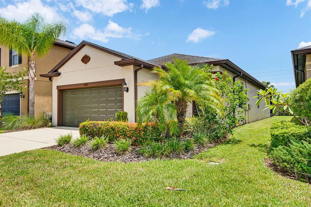 A single-story house with a tan stucco exterior, a dark garage door, and front landscaping — 1407 CUSTER BAYOU STREET, RUSKIN, FL
