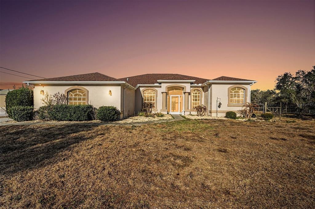 A single-story stucco house with a brown shingled roof stands under a sunset sky — 16031 DONNEY MOOR LANE, SPRING HILL, FL