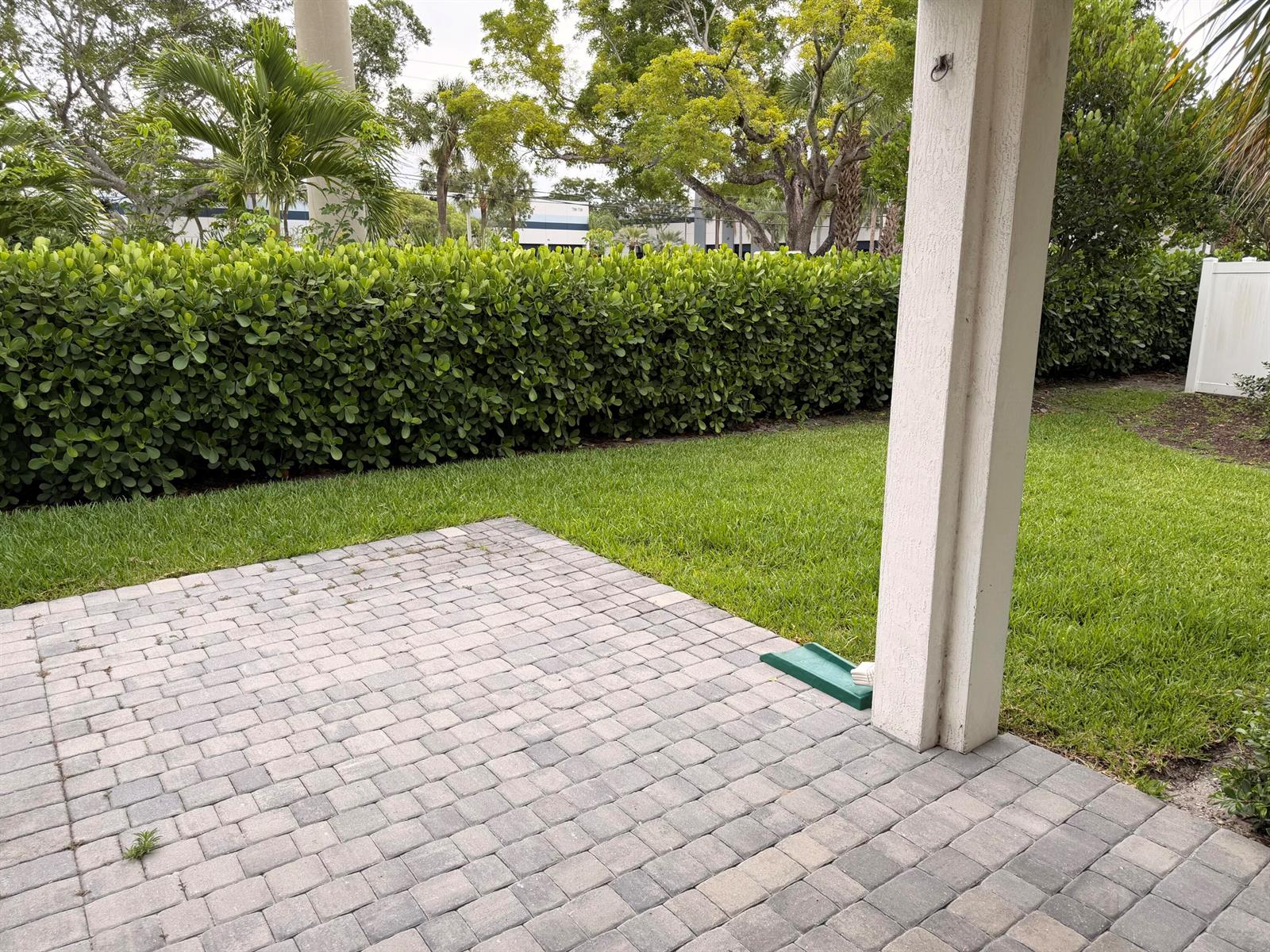 A paved patio overlooks a green lawn bordered by a tall hedge and trees — 562 Parsons Way, Deerfield Beach FL 33442, DEERFIELD BEACH, FL