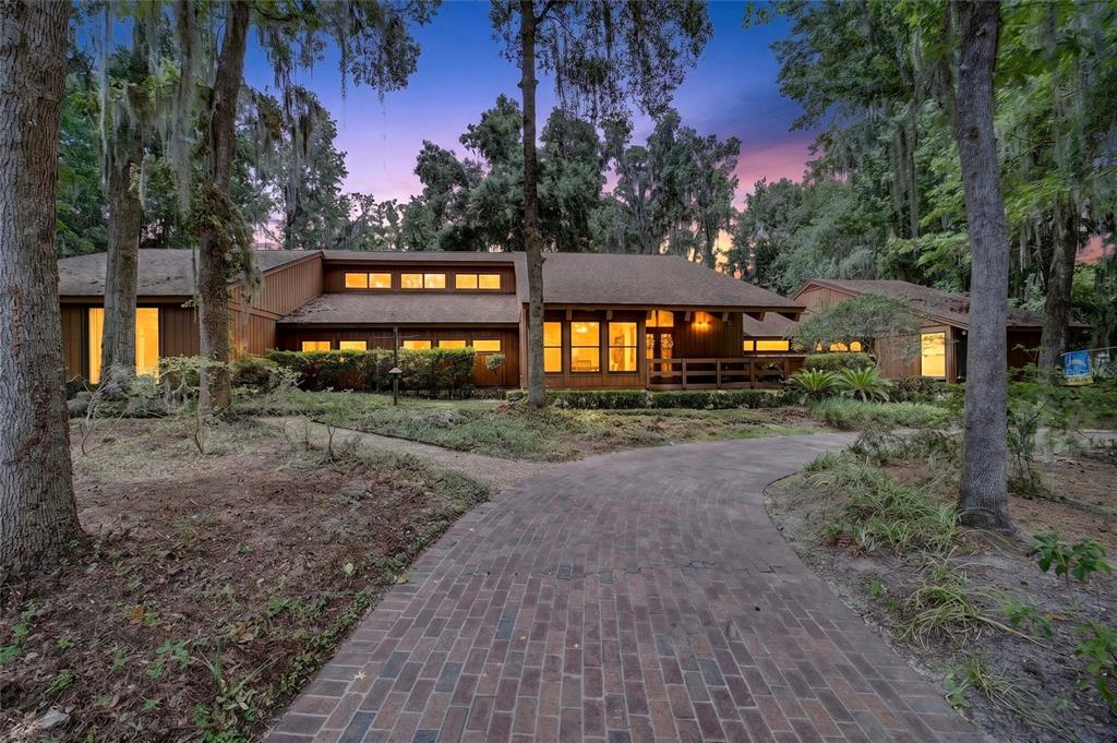 A brown wood-sided house with a brick driveway sits among trees at dusk — 2411 SE 15TH STREET, OCALA, FL
