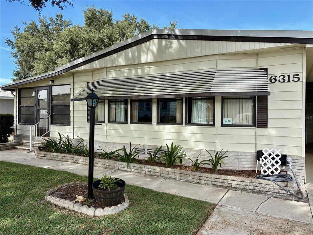 The exterior of a light-colored manufactured home features a brick planter and metal window awnings — 6315 GROVE POINTE DRIVE SE, WINTER HAVEN, FL