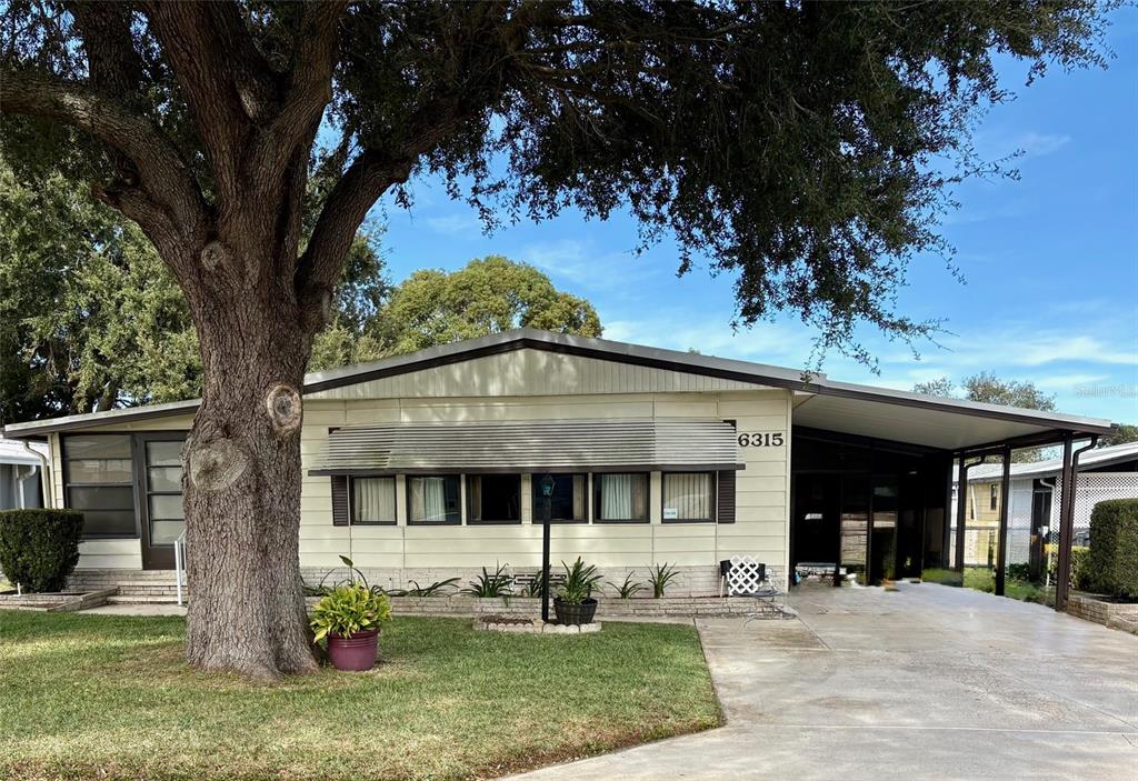 A single-story mobile home with light siding, a large shade tree, and a carport — 6315 GROVE POINTE DRIVE SE, WINTER HAVEN, FL