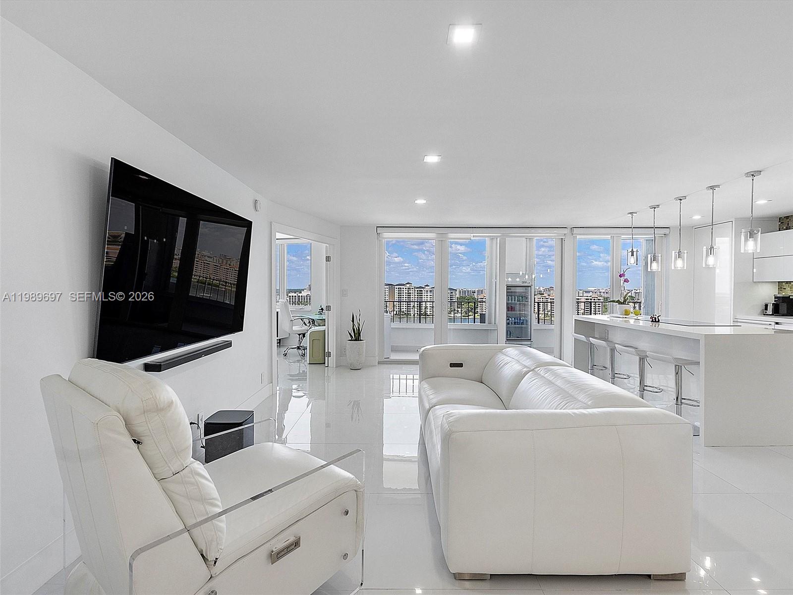 A modern white living room features a large television, leather seating, and a kitchen island — 550 S Ocean Blvd # 1509, Boca Raton FL 33432, BOCA RATON, FL