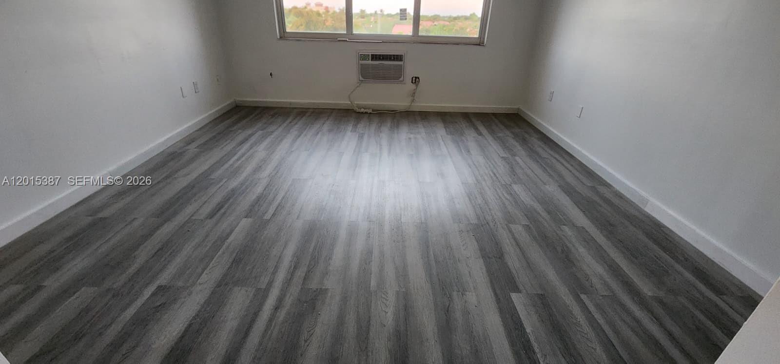An empty room features gray wood-look flooring, white walls, and a window air conditioning unit — 6930 Miami Gardens Dr # 512A, Hialeah FL 33015, HIALEAH, FL