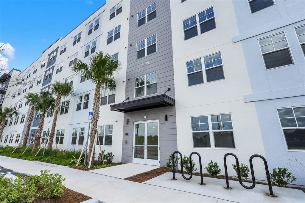 The exterior of a modern multi-story apartment building features a glass entrance and bike racks — 5579 MILLENIA PARK BOULEVARD UNIT 508, ORLANDO, FL
