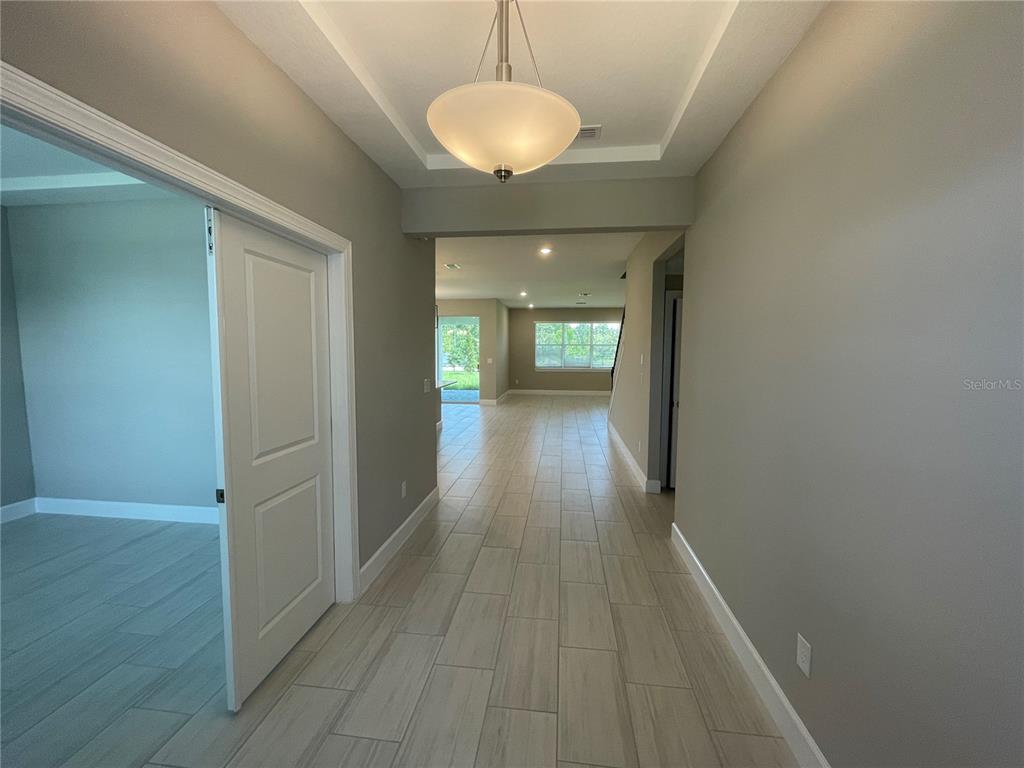 A hallway with light-colored tile flooring leads toward an open living area with large windows — 34286 EVERGREEN HILL COURT, WESLEY CHAPEL, FL