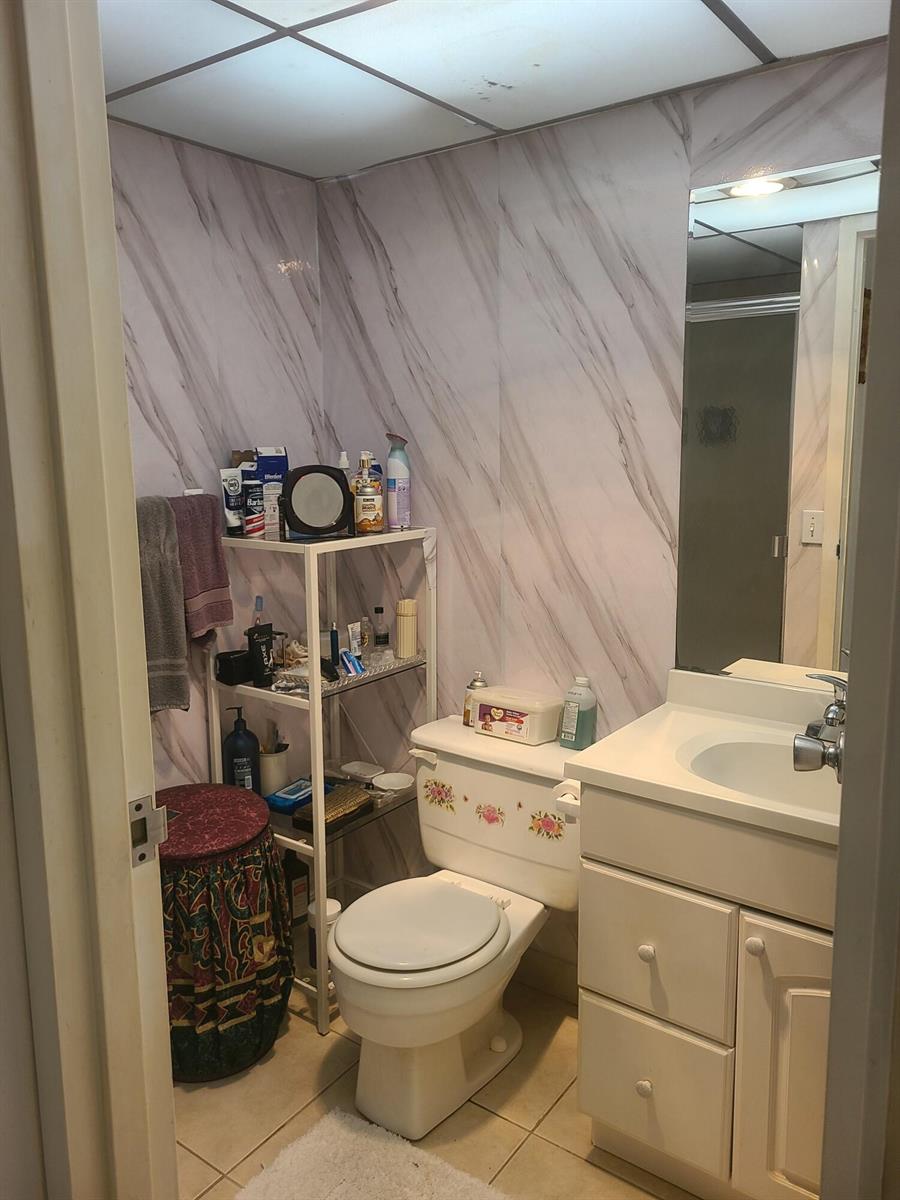 A small bathroom features marble-patterned walls, a white vanity, a toilet, and a metal shelving unit — 257 Brittany F # 257, Delray Beach FL 33446, DELRAY BEACH, FL