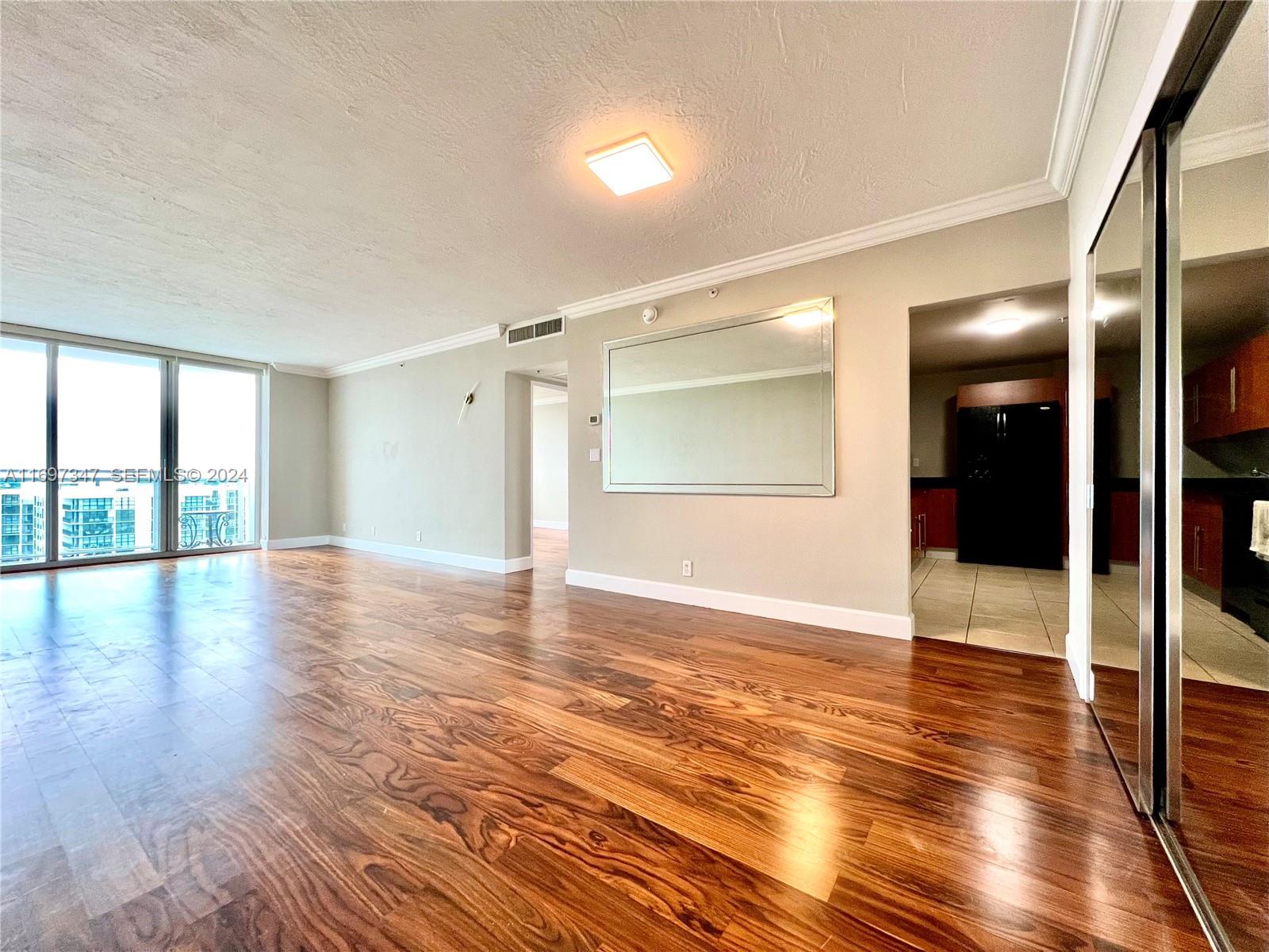 An empty living room features wood flooring, a large wall mirror, and floor-to-ceiling windows — 3000 S Ocean Dr # 1012, Hollywood FL 33019, HOLLYWOOD, FL