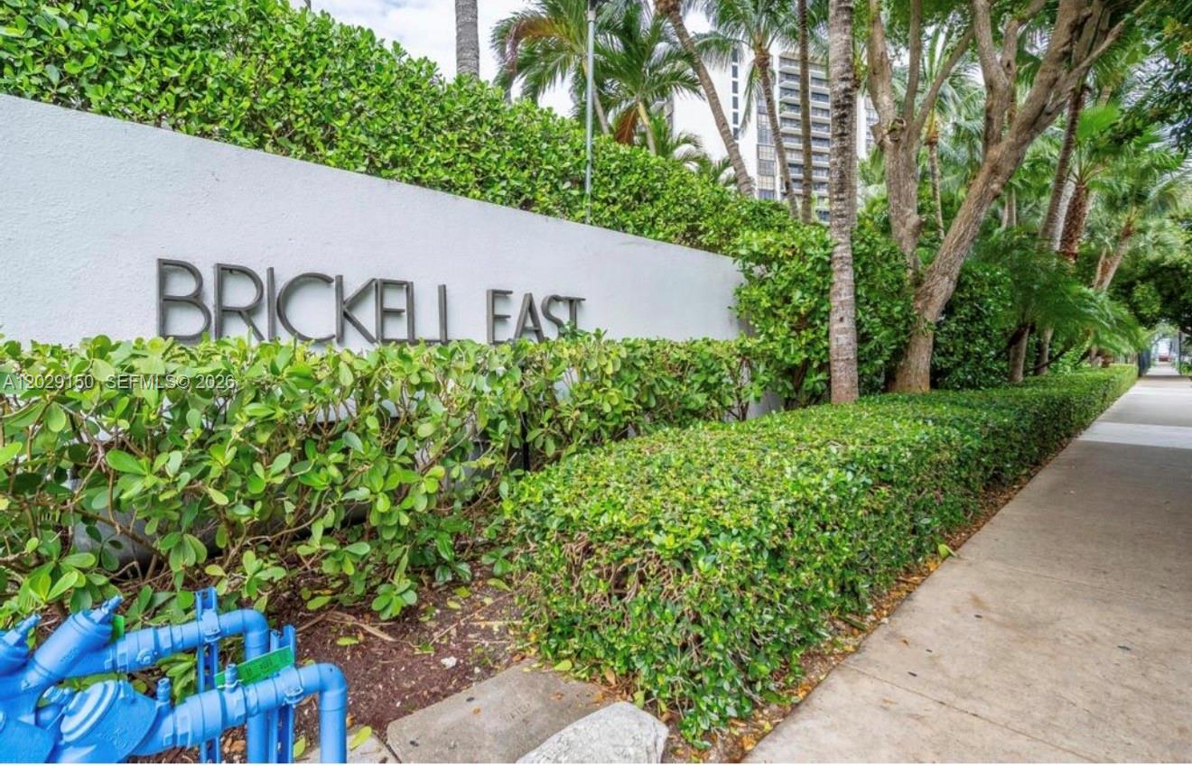 A white wall with the text Brickell East stands behind a hedge next to a sidewalk — 151 SE 15th Rd # 502, Miami FL 33129, MIAMI, FL