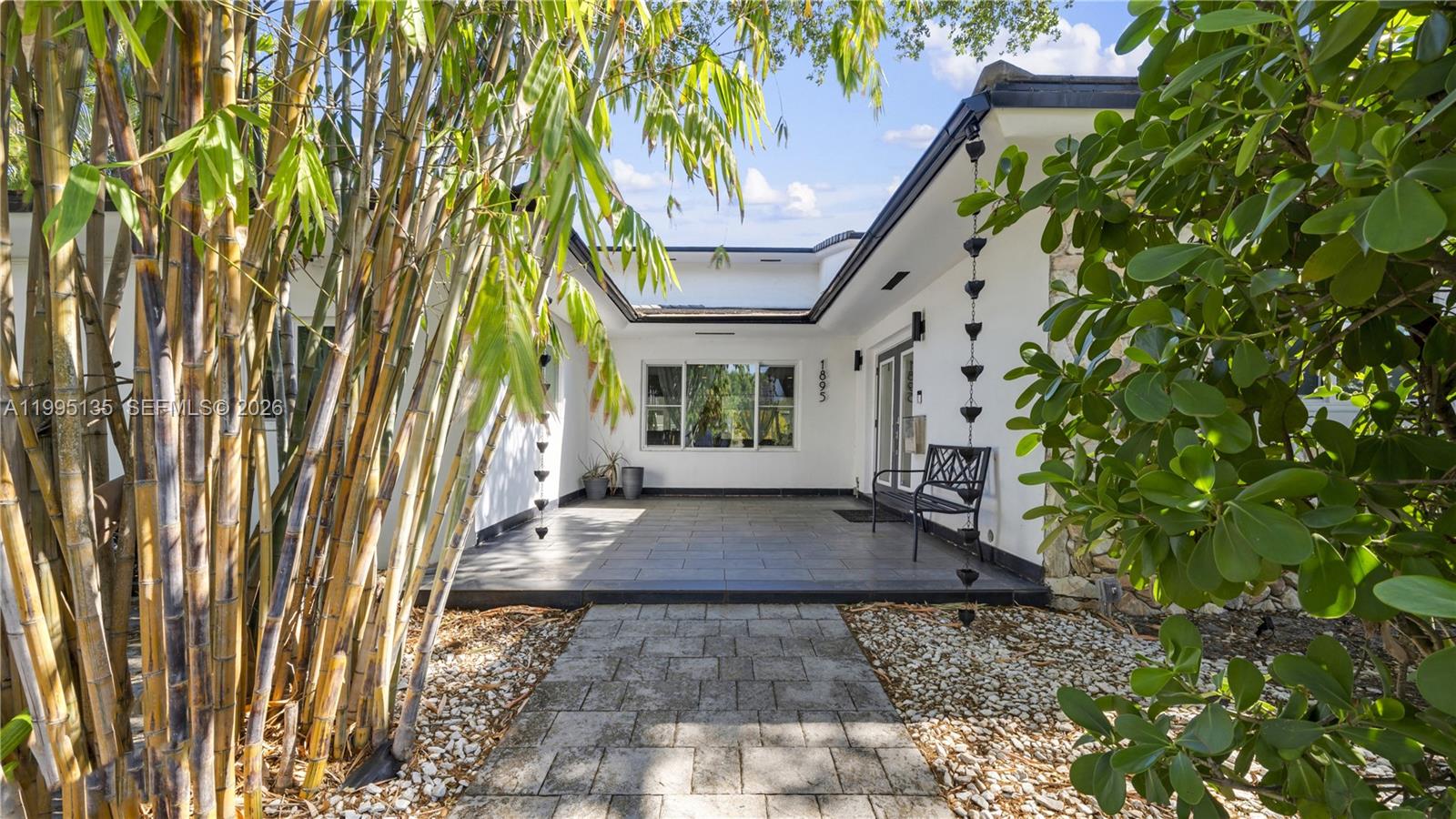 A paved walkway leads to a covered patio entrance with white walls and bamboo landscaping — 1895 NE 118th Rd, North Miami FL 33181, NORTH MIAMI, FL